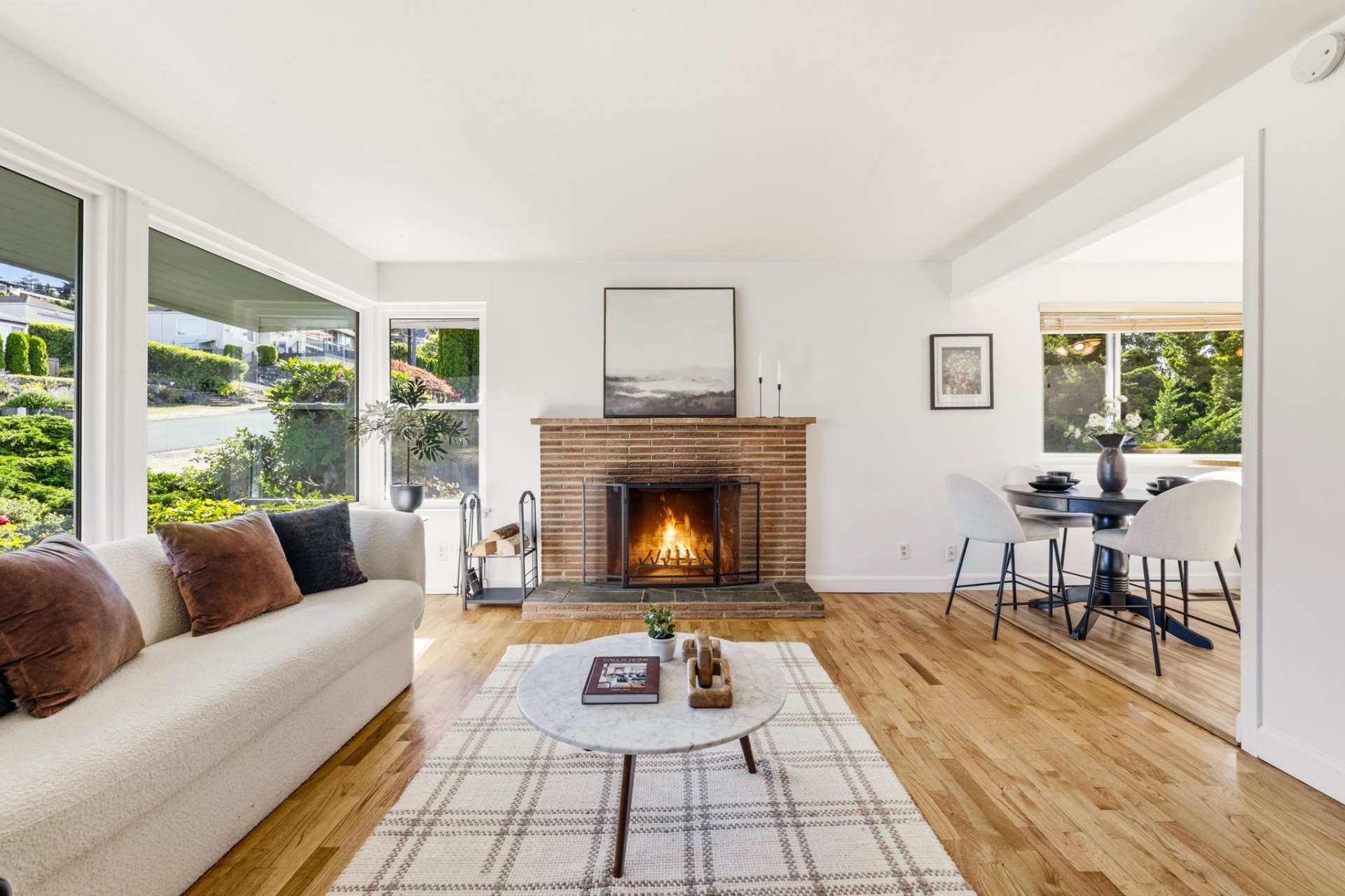 Bright living room with hardwood floors, a lit brick fireplace, a marble coffee table, and a dining nook filled with natural light