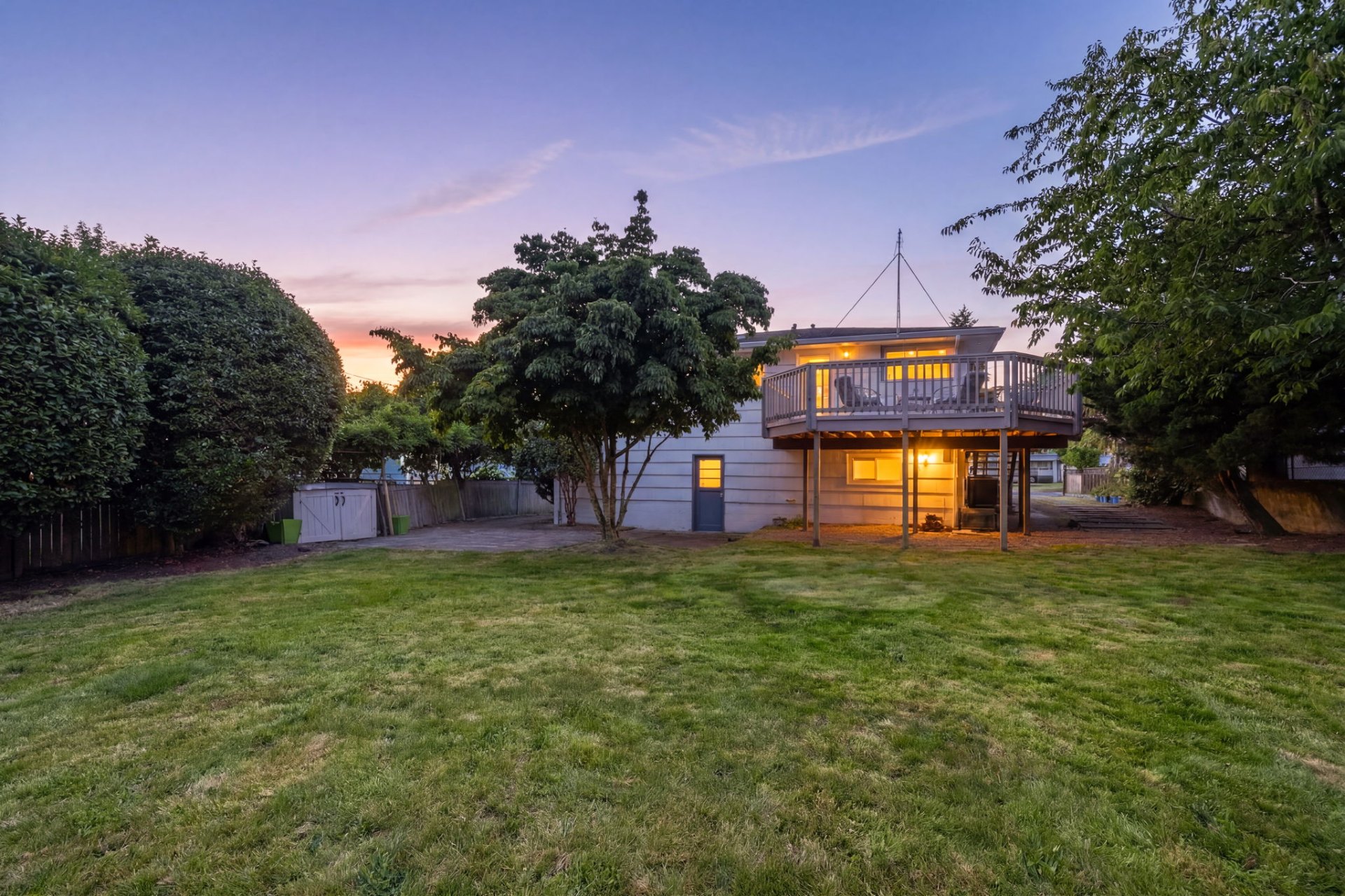 Spacious backyard at twilight with a large lawn, elevated deck, and the rear of the home warmly lit